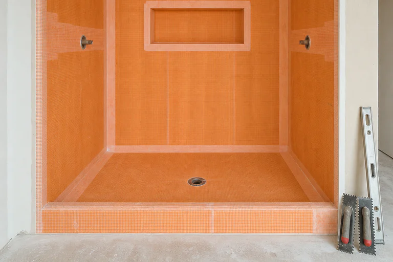 Shower waterproofing stage with a membraned sloped pan, banded corners, a wrapped curb, and trowels staged on a drop cloth