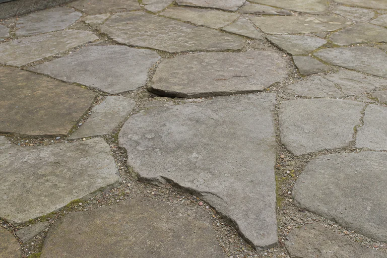 Weathered flagstone patio with localized settlement, open aggregate joints, and moss at a shaded edge
