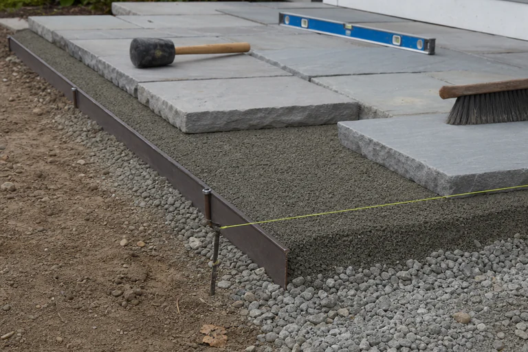 Flagstone installation showing compacted aggregate, bedding layer, edge restraint, fitted stones, string line, level, and mallet