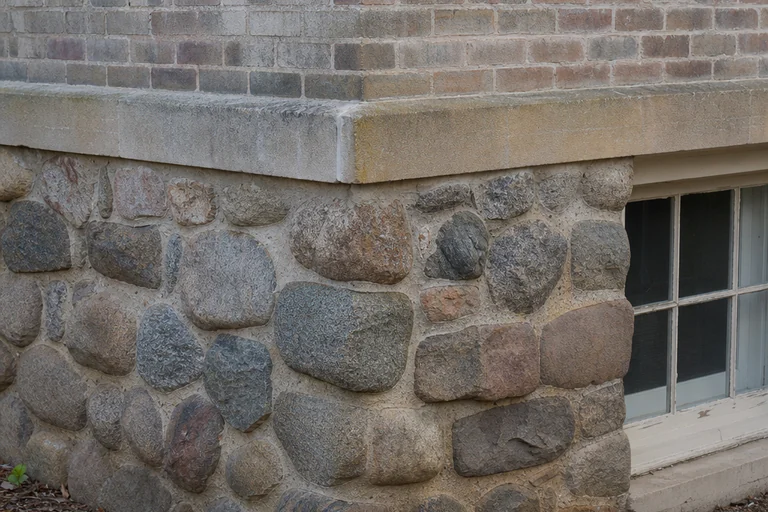 Rounded fieldstone foundation below a limestone water table and weathered brick beside a basement window